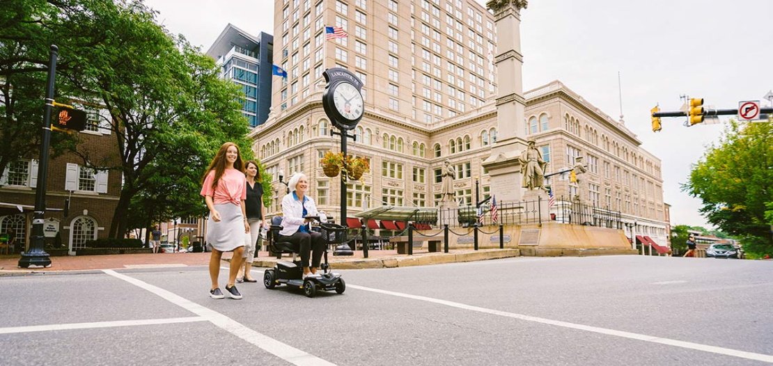 An elderly woman crosses the street with a four-wheeled scooter next to a woman and young woman.