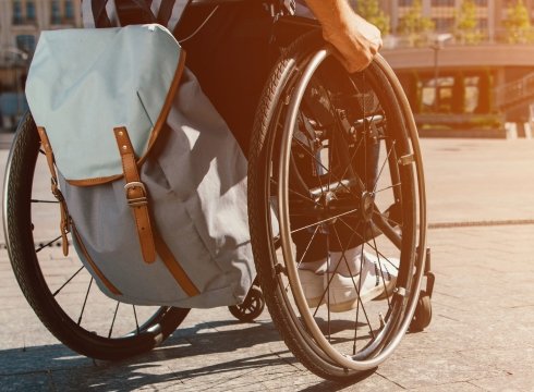The Backside Of A Wheelchair With A Person’S Hands On The Wheels, Pushing Across A Large Town Square In Orange Lighting