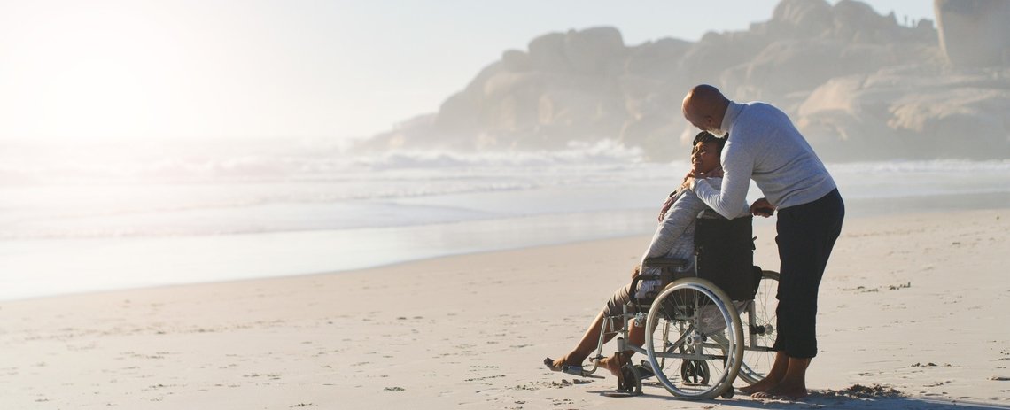 A Man Stands By A Woman In A Manual Wheelchair On A Sandy Beach