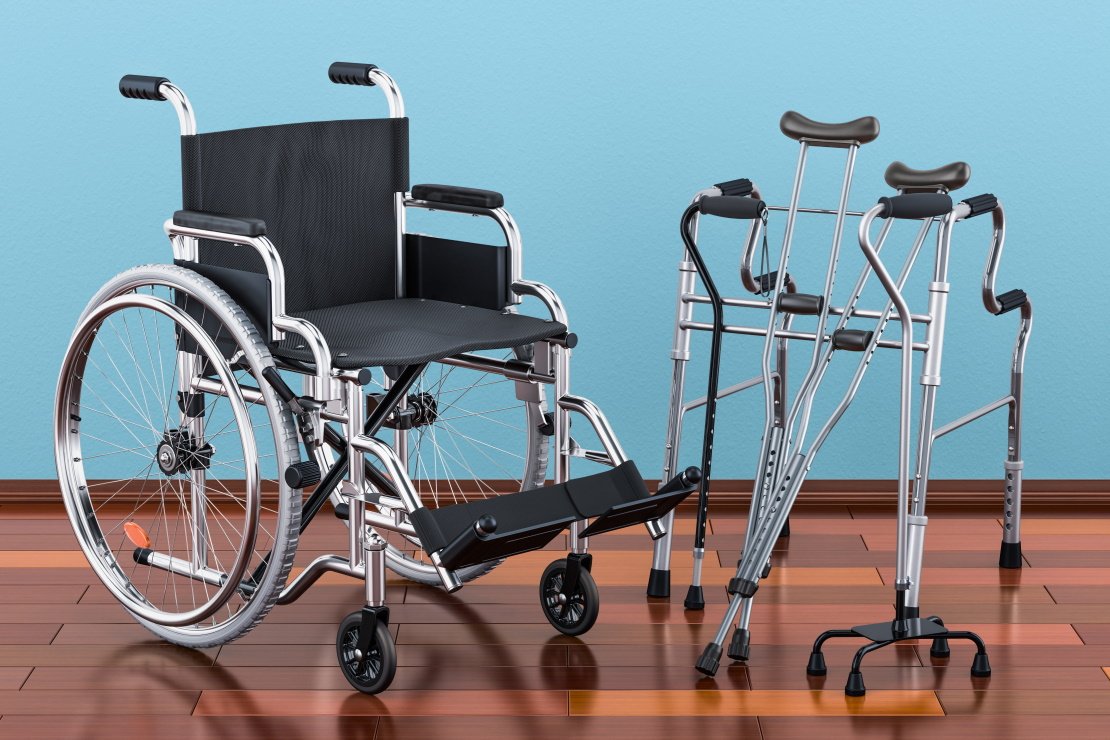 A wheelchair, a rollator, two canes and crutches on a brown floor, blue wall of background.
