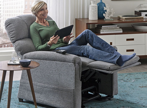 A Smiling Woman Sitting On A Gray Golden Technologies Power Lift Recliner Next To a Table With A Glass Of Water On It