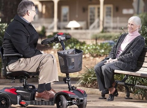 An elderly man on a mobility scooter talks to an elderly woman sitting on a park bench